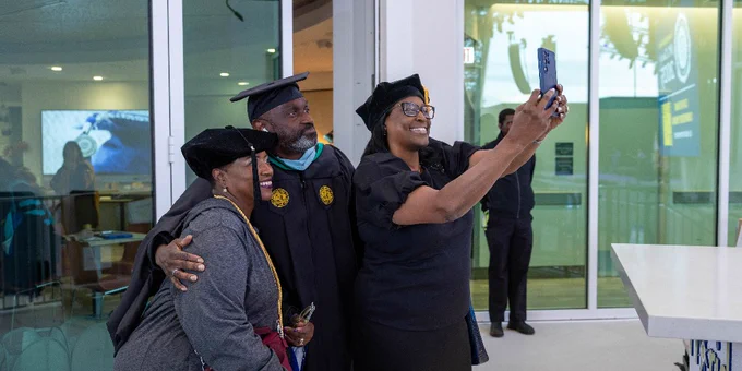 SPC President Dr. Tonjua Williams and two other individuals in academic regalia take a selfie together. They are standing outside a building with large windows. One is holding a phone for the selfie. 