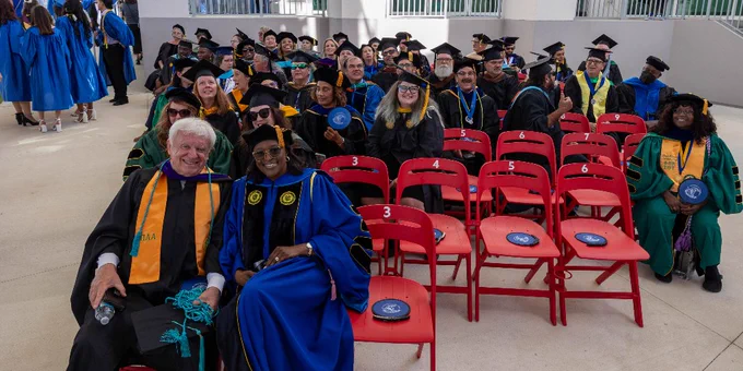 A large group of faculty in academic regalia seated on red chairs and smiling at a graduation ceremony.