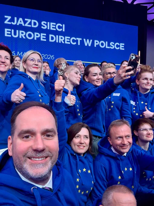 Zdjęcie przedstawiające wiele uśmiechniętych osób w tym autora posta i komisarza Piotra Serafina na podium w niebieskich bluzach z flagą UE
