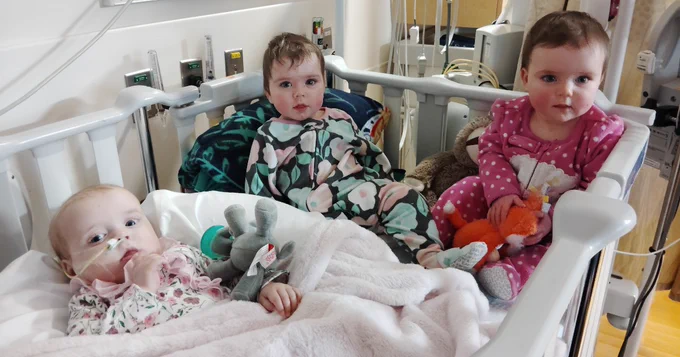 Three young children are sitting in a hospital crib. They are surrounded by stuffed animals and wear colorful outfits. Medical equipment is visible in the background.