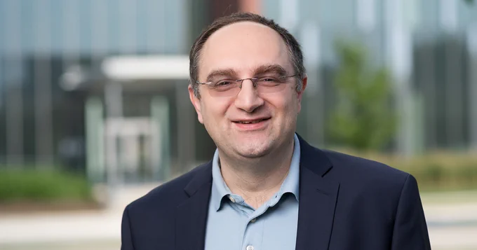 A portrait of Tillman Kubis. A building with large glass windows is blurred in the background.