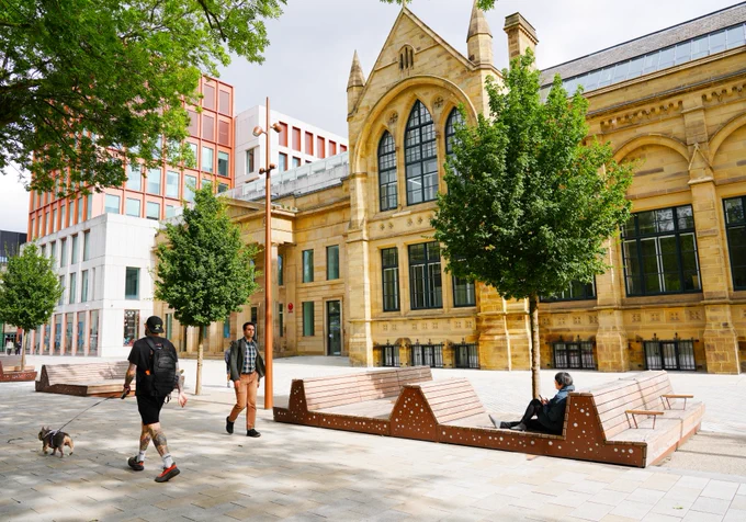 A street scene in front of the Manchester Metropolitan University building, featuring a historic, yellow-toned structure with arched windows. Several people and a small dog are walking or sitting on modern wooden benches. Trees line the paved area, and a contemporary building is visible in the background.