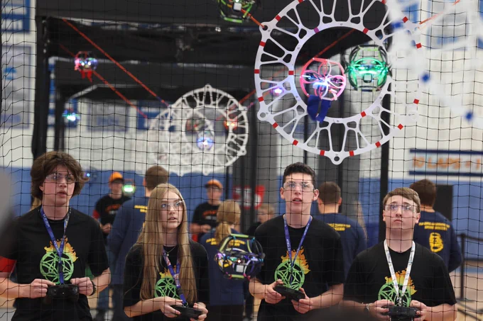 Students wearing safety glasses pilot glowing drone soccer drones inside a netted arena, aiming to fly a caged drone through a circular goal during a competitive match.