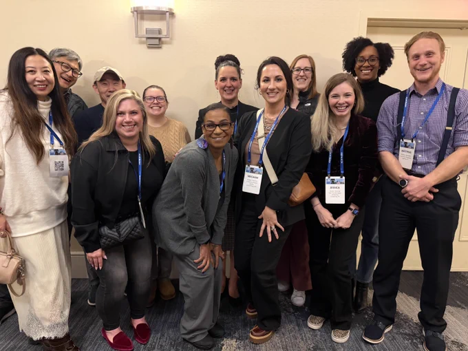 A group of people smiling and posing together at an indoor event. Most are wearing conference name tags and blue lanyards. They are casually dressed and standing against a light-colored wall, with a light fixture above them.
