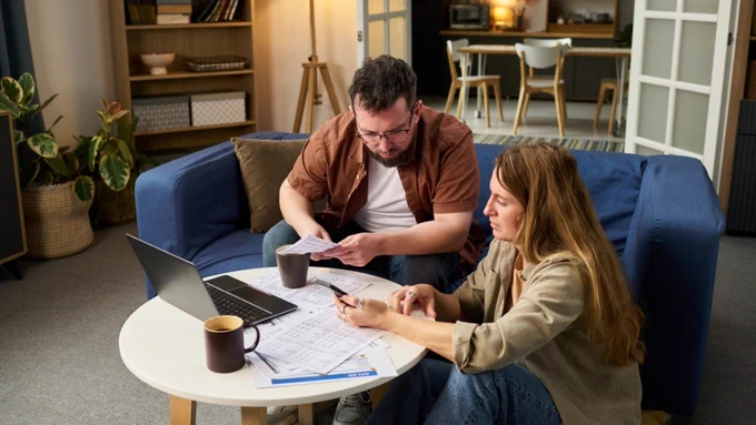 Two people sitting in a living room, reviewing documents at a small round table. A laptop and coffee mugs are also on the table. In the background, there are bookshelves, plants, and a dining area.