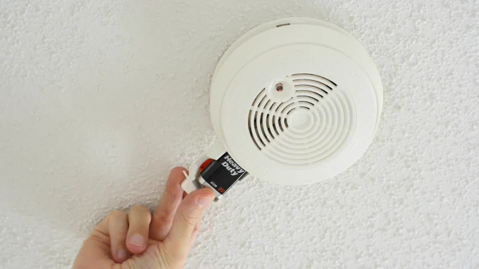 Hand replacing a 9V battery in a white ceiling smoke detector mounted on a textured surface.