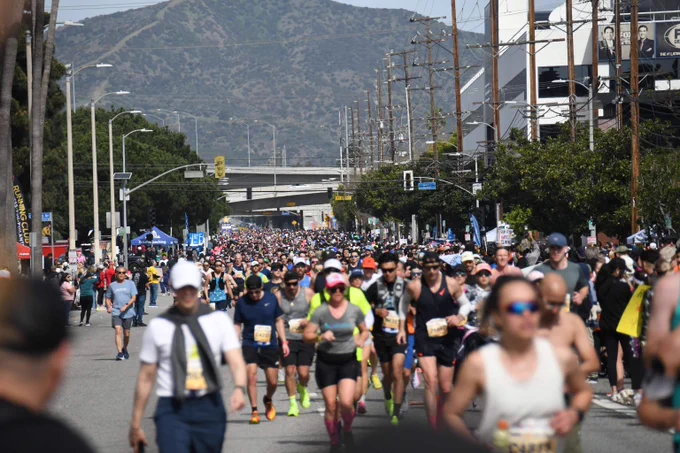 Large crowd of runners participating in a marathon on a city street with mountains in the background under clear skies.