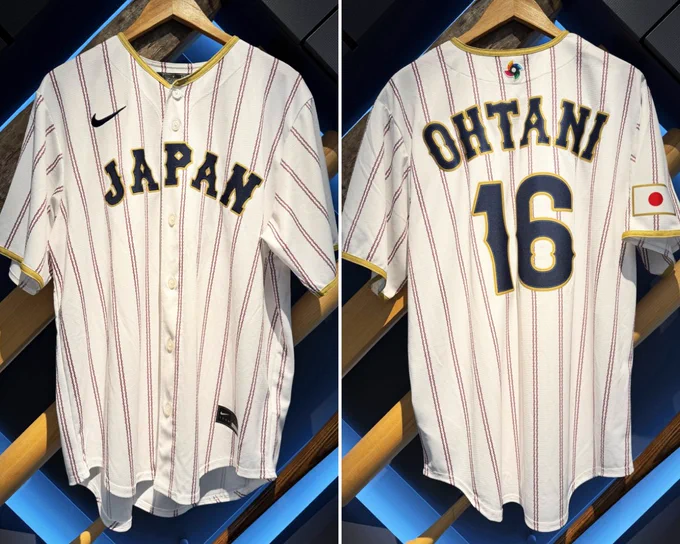 Left, the front of a white Team Japan uniform with navy lettering, gold trim and red pinstripes. Right, the back of a Shohei Ohtani white Team Japan uniform with navy lettering, gold trim and red pinstripes.