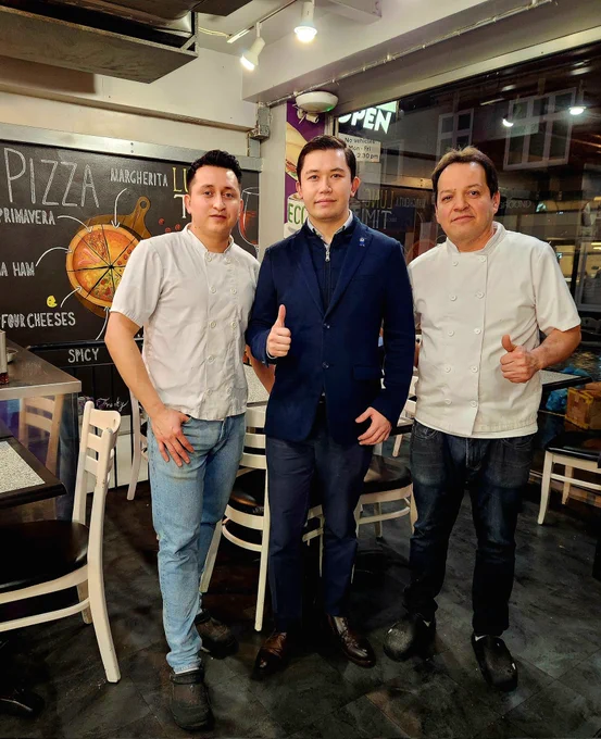 Three men standing side by side in a pizzeria, two of them wearing a chef's. 