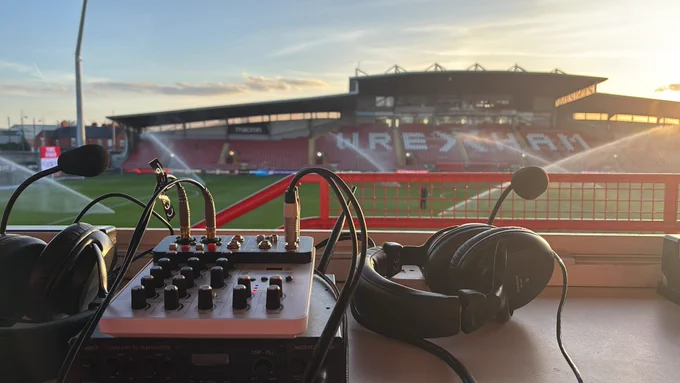 Audio description commentary kit placed in the media row of the top tier of the Wrexham lager stand. The macron stand bathed in hazy sunshine can be seen in the background.