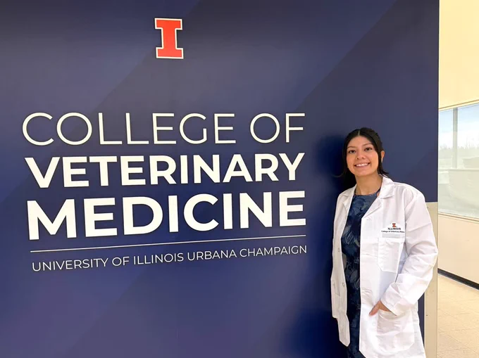 A Texas A&M University-Corpus Christi alumna wearing a white coat stands smiling beside a wall displaying the "College of Veterinary Medicine, University of Illinois Urbana-Champaign" sign.