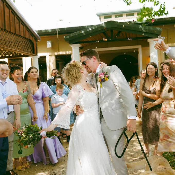 A joyful wedding scene with a Texas A&M University-Corpus Christi alumnus in a light suit and another in a white dress sharing a kiss among cheering guests. Some guests hold flower bouquets, and a dog is barely visible at the bottom. The setting is outdoors with a building in the background.