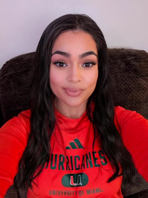 A Texas A&M University-Corpus Christi alumna with long dark hair is sitting on a dark brown chair, wearing a red University of Miami Hurricanes shirt. The shirt has the Adidas logo above the lettering. The background is a light gray wall.