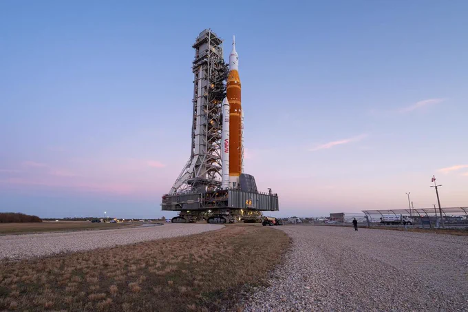 NASA’s Artemis II SLS (Space Launch System) rocket and Orion spacecraft roll back to the Vehicle Assembly Building at NASA’s Kennedy Space Center in Florida, Feb. 25, 2026. In the foreground, dry grass and two gravel paths outline the route for the Crawler-Transporter. The sky above is blue and pink with wispy clouds.
Photo Credit: (NASA/John Kraus) 
