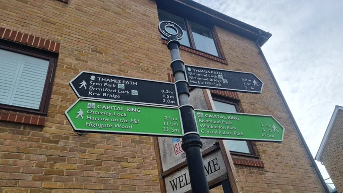 A shared finger post at Isleworth with the Thames Path signs in black and the Capital Ring in lime green.  In the background is a small apartment block.