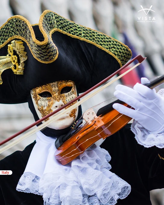A person wearing an ornate Venetian mask and costume plays a violin. They are dressed in a black and gold hat, with white lace ruffles, embodying the Venetian carnival tradition. The background is blurred but suggests a historic European setting.