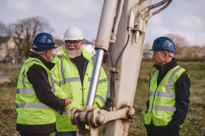 3 people stand next to the arm of a digger