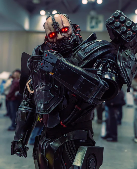 Cosplayer dressed in a realistic Adam Smasher outfit poses with one arm raised across their chest, their hand closed in a tight fist. Glowing red eyes are lit up in the middle of a pallid face. The cosplayer wears a complex bodysuit resembling a full cyborg conversion. In the background, other convention attendees are visible.