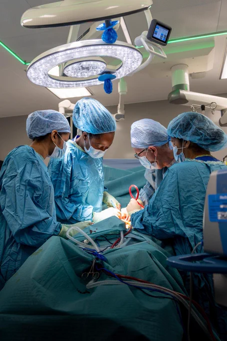 Staff working during the penile implant surgery. They are wearing full personal protective equipment, including scrubs, aprons, surgical caps and face masks. The patient on the operating table is covered by surgical drapes.
