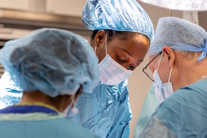 A candid, close up photo of Consultant Urological Surgeon and Andrologist, Elizabeth Osinibi, during surgery. She is wearing full PPE including blue scrubs, a surgical cap and face mask. She is looking down at the operating table, which is not in view in the photo, while two colleagues in the foreground assist with the procedure.