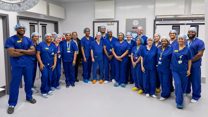 Around 20 staff involved in the procedures pose for a group photo in the empty operating theatre. Most are wearing blue scrubs.