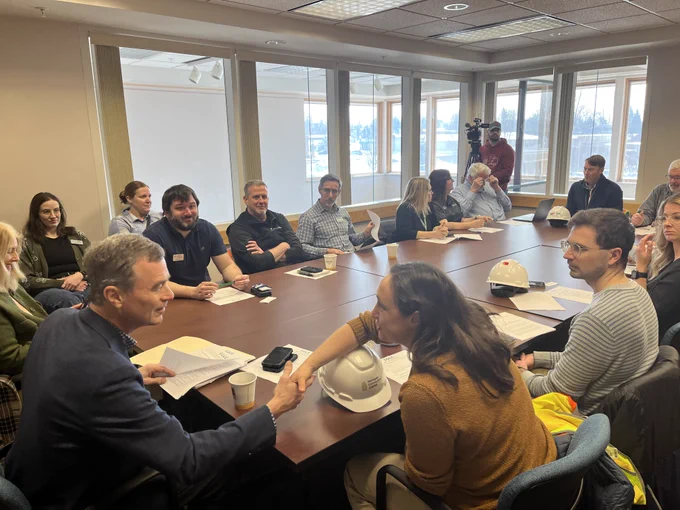 State and local leaders meet with EGLE staff around a large conference table. Papers, notebooks, and hard hats are on the table, and a camera operator records from the back of the room. Large windows provide natural light.