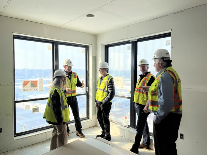 GLE Director Phil Roos, staff, and leaders wearing hard hats and safety vests stand inside a corner room of the developing building of Cadillac Lofts, discussing progress. Large windows behind them show the outdoor construction area and winter landscape.