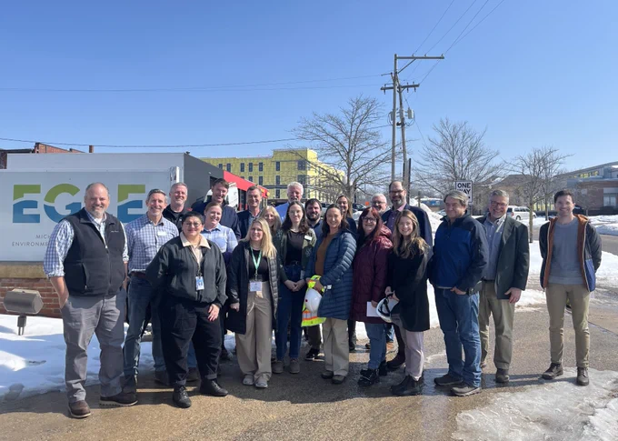 A group of EGLE staff and state and local leaders stand together outdoors in front of an EGLE sign on a sunny winter day. Snow is on the ground and construction buildings are visible in the background.