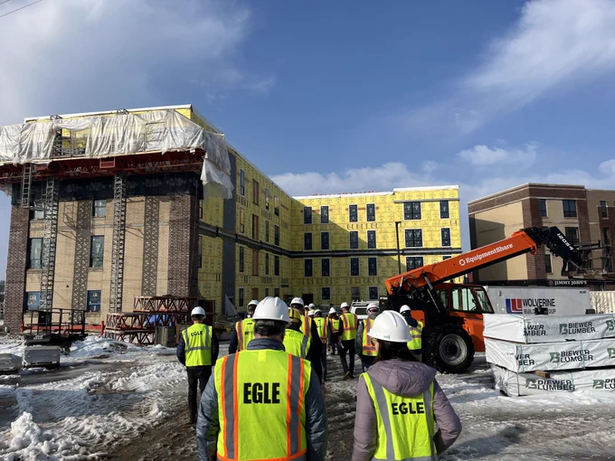 Local leaders and EGLE staff wearing safety vests and hard hats walk toward Cadillac Lofts, a large multi‑story building under construction. The structure is covered in yellow sheathing, and construction equipment sits nearby on the snowy ground.