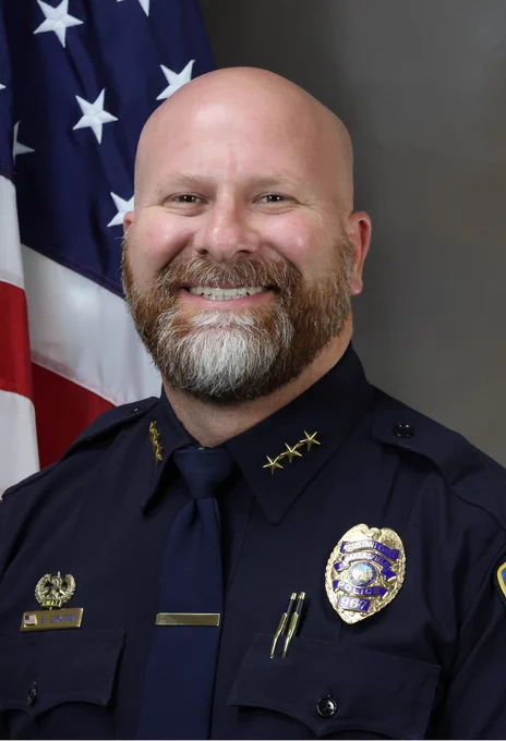 Bakersfield Police Department Chief Stratton in his Class A police uniform stands smiling in front of a United States flag. The uniform includes a badge and shoulder stars.