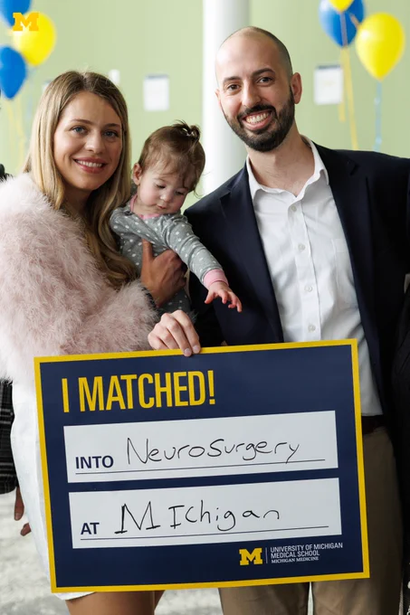 A U-M Medical School Student taking a photo with another adult and child with a sign that says they matched into neurosurgery at Michigan.