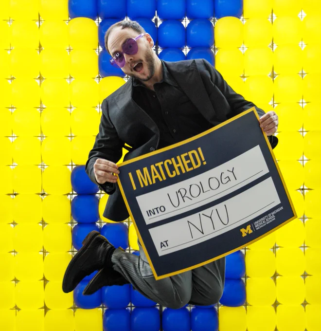 A U-M Medical School student jumping with a sign that says they matched into urology at NYU.