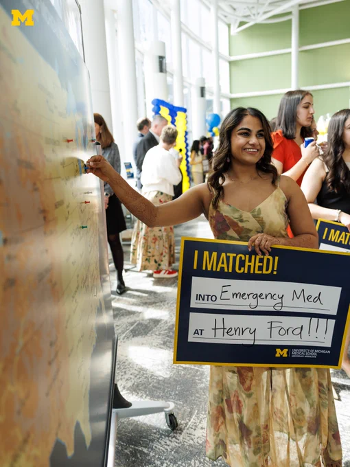 A U-M Medical School student smiling while putting a pin on a map and holding a sign that says they matched into emergency medicine at Henry Ford.