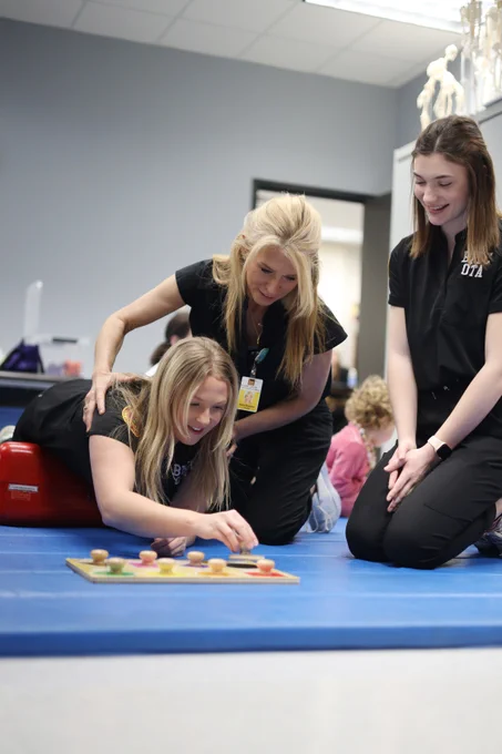 Two people are assisting another in an occupational therapy setting. One is laying on a red bolster, engaging with a colorful pegboard. The room has therapy equipment and other individuals in the background.