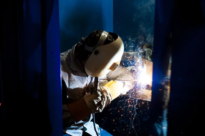 A person is welding metal indoors. They are wearing protective gear, including a helmet and gloves. Bright sparks and a glow illuminate the area as the welding torch is in use.