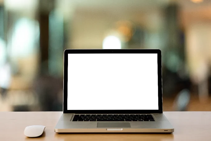 A laptop with a blank white screen sits on a wooden desk, with a wireless mouse beside it, against a softly blurred indoor background.