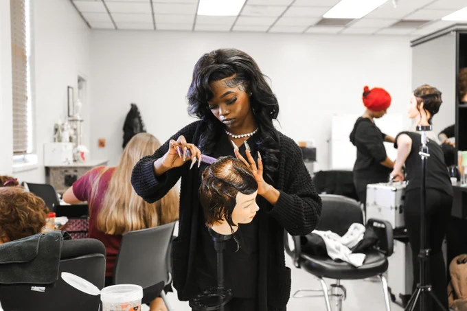In a salon setting, an individual styles a mannequin's hair with focus and precision. Others work on different tasks in the background. 