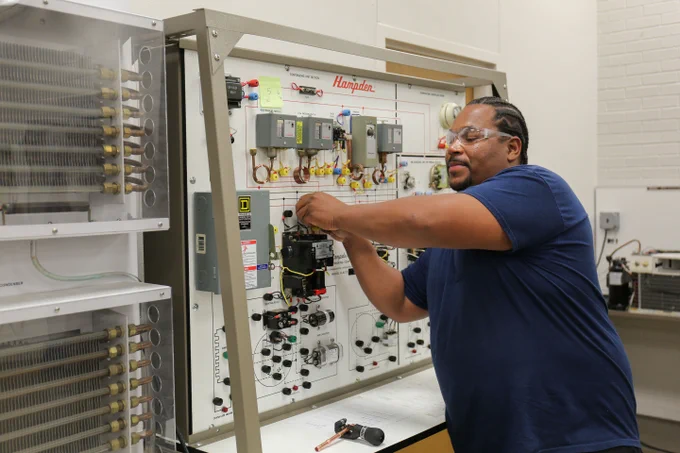 A person is working on a training board in a lab. They are adjusting wires and components, and wearing safety glasses.