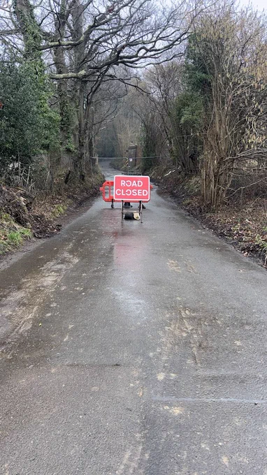 A 'Road Closed' sign sits in the middle of a tree lined road, the road is Broom Lane.
Behind the sign a can be seen a gap where the old bridge once carried the Spa Valley Railway over Broom Lane