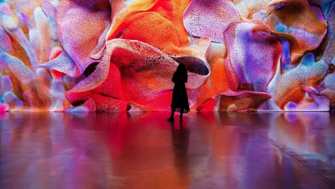 A viewer experiencing a Refik Anadol AI-generated data sculpture. The vibrant, organic-looking digital waves fill the room with light, reflecting off the gallery's glossy surface.