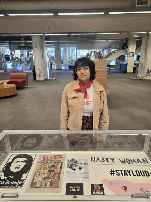 Woman with dark, short hair in camel colored coat with red neck bandana standing behind the glass case with materials. 
