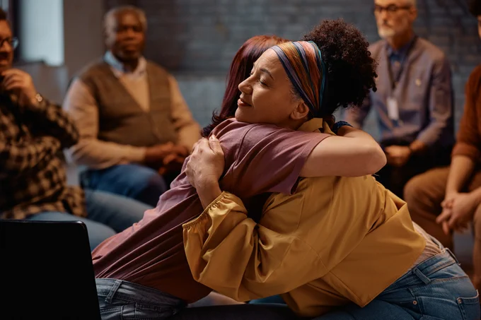 Two women warmly embrace during a group therapy session with others seated in the background.