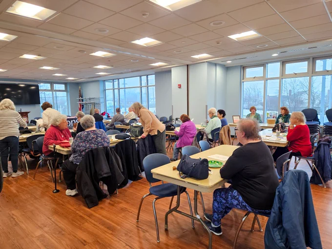 In a center, tables of older adults sit together and are visited by staff members walking around the room.