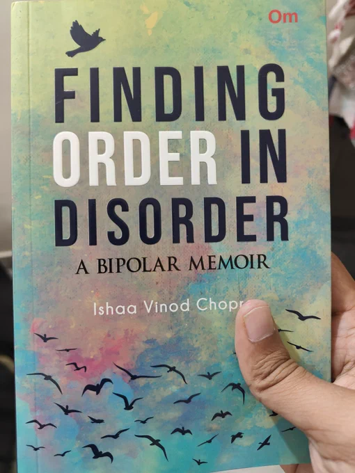 A close-up photo of the front cover of the book Finding Order in Disorder: A Bipolar Memoir by Ishaa Vino Chopra, published by Om Books International. The cover features a teal-to-pink gradient background with abstract silhouettes of several black birds flying across it. Bold white text reads "FINDING ORDER IN DISORDER" at the top and "A BIPOLAR MEMOIR" below, with the author's name in smaller black text at the bottom. The book is held in a hand visible at the bottom edge, with the publisher's red "Om" logo in the top right corner. 