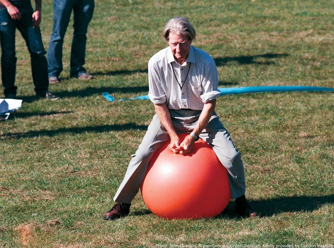 John Gurdon sitting on a large red exercise ball in a grassy field, appearing to balance. Others stand in the background.