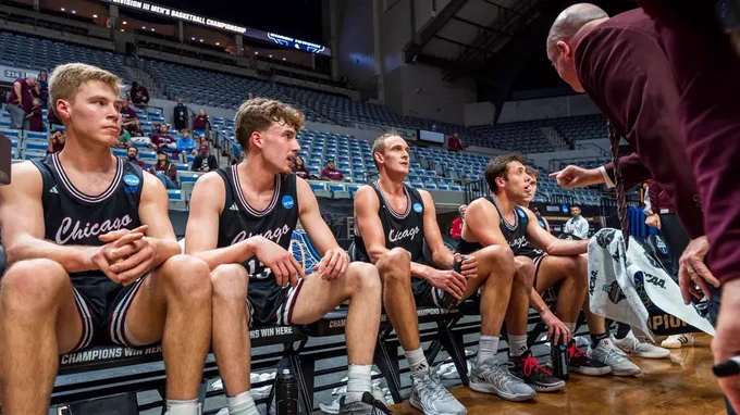 A coach points and speaks emphatically to four basketball players in black Chicago jerseys on the bench. The players are looking at the coach with varying expressions. Text on the bench reads \"CHAMPIONS WIN HERE\".