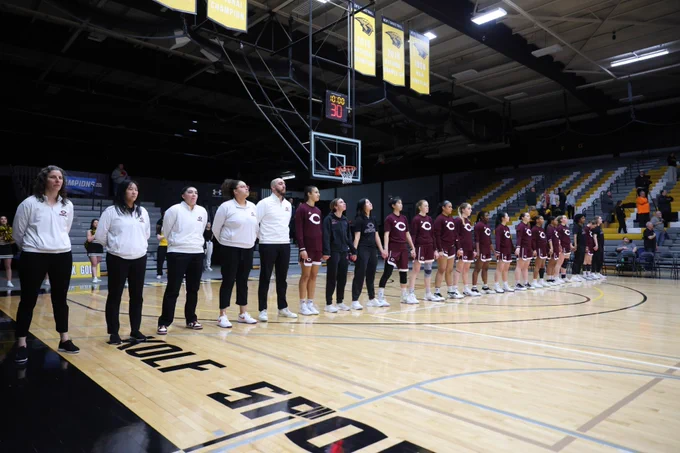 A basketball team stands on court during the national anthem. The scoreboard reads text@1. Banners hang above, celebrating past wins. Team wears maroon uniforms with white trim; coaches in white tops and black pants.
