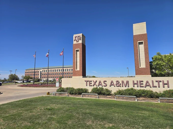 Texas A&M Health campus under a blue sky with the American, Texas and Texas A&M flags flying above landscaping featuring pink flowers