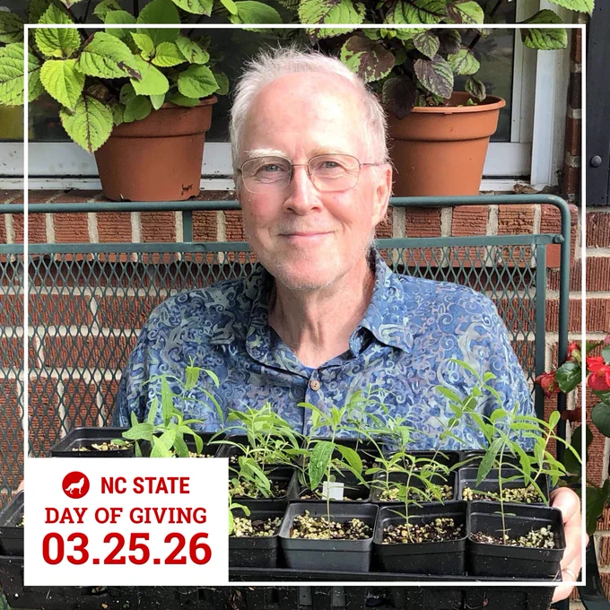 NC State Extension Master Gardener volunteer John Bowen holding a tray of young green plants in small pots, sitting outdoors with leafy plants in the background.