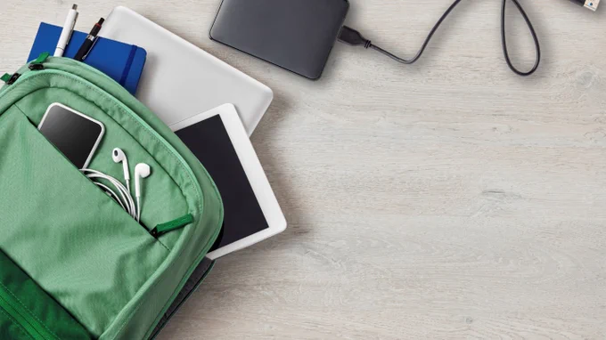 A green backpack on a light wooden surface is partially open, revealing a smartphone, earphones, pens, a closed blue notebook, and a white tablet. Nearby, a silver laptop and black external hard drive connected via a cable are arranged neatly.